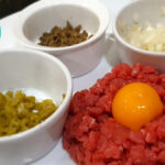Tartare de boeuf au couteau. Image showing finely hand-chopped fillet steak with an egg yolk in the centre and small bowls containing the seasonings on the side - one with finely chopped gherkins, one with finely chopped capers and the third with finely chopped onions.
