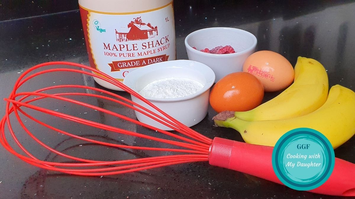 Ingredients for Banana Pancakes with Logo. Image shows 2 bananas, 2 eggs, some flour in a small bowl, a few raspberries in a small bowl alongside a red whisk and a bottle of maple syrup on a black worktop.