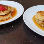 Pncakes with Logo. Image shows two plates on a black worktop which each have 2 pancakes on covered in maple syrup. The left hand plate of pancakes has a few raspberries on top.