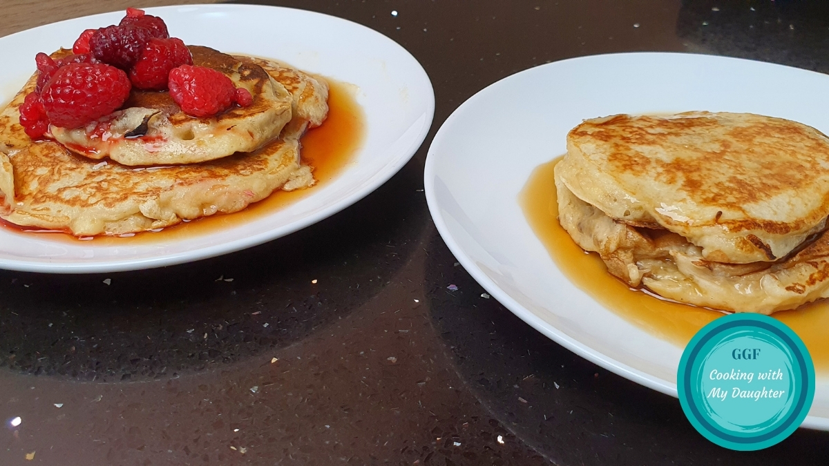 Pncakes with Logo. Image shows two plates on a black worktop which each have 2 pancakes on covered in maple syrup. The left hand plate of pancakes has a few raspberries on top.